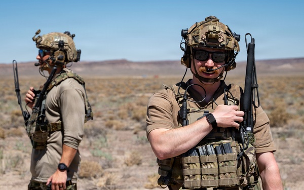 Two armed military or special operations personnel are standing in an arid desert environment with sparse vegetation. Both individuals are equipped with tactical gear including helmets, plate carriers, weapons, and communication equipment. The setting appears to be a training exercise or operational deployment in a desert region.