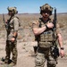 Two armed military or special operations personnel are standing in an arid desert environment with sparse vegetation. Both individuals are equipped with tactical gear including helmets, plate carriers, weapons, and communication equipment. The setting appears to be a training exercise or operational deployment in a desert region.