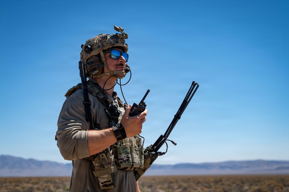 A military operator in full tactical gear stands in an arid desert environment under a clear blue sky. He is holding a radio communications device with multiple antennas and appears to be monitoring or coordinating communications. The background reveals a flat desert landscape with distant mountain ridgelines.