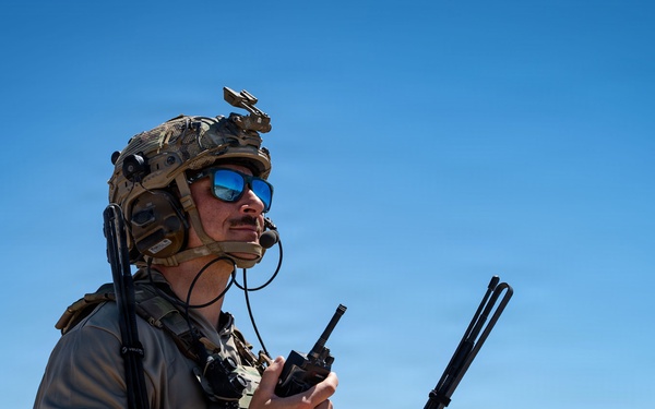 A military operator in full tactical gear stands in an arid desert environment under a clear blue sky. He is holding a radio communications device with multiple antennas and appears to be monitoring or coordinating communications. The background reveals a flat desert landscape with distant mountain ridgelines.