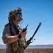 A military operator in full tactical gear stands in an arid desert environment under a clear blue sky. He is holding a radio communications device with multiple antennas and appears to be monitoring or coordinating communications. The background reveals a flat desert landscape with distant mountain ridgelines.