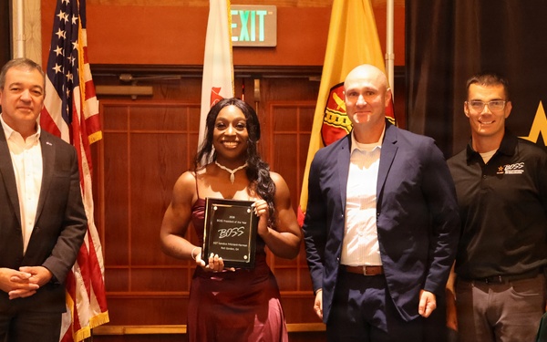 Four individuals pose for a formal photo at a BOSS (Better Opportunities for Single Soldiers) award ceremony. A woman in a burgundy evening gown holds a black plaque award while standing between three men in formal attire. The event appears to be a military-affiliated recognition ceremony with flags and branded banners visible in the background.