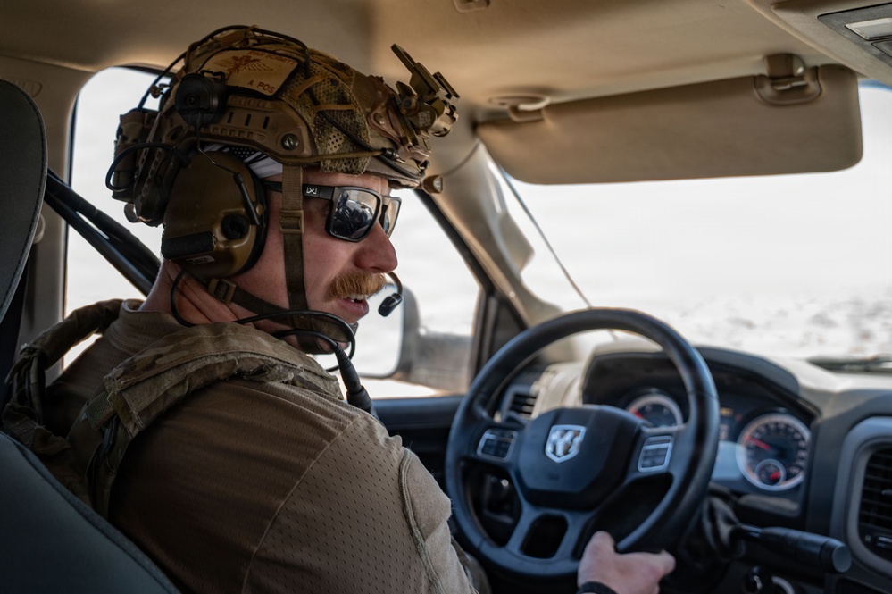 A military or tactical operator is driving a Ram pickup truck through what appears to be an arid, desert-like environment. The individual is wearing full tactical gear including a combat helmet with mounted accessories, communications headset with boom microphone, sunglasses, and a plate carrier or tactical vest. The vehicle interior and steering wheel are clearly visible, with the Ram logo prominently displayed on the steering wheel.