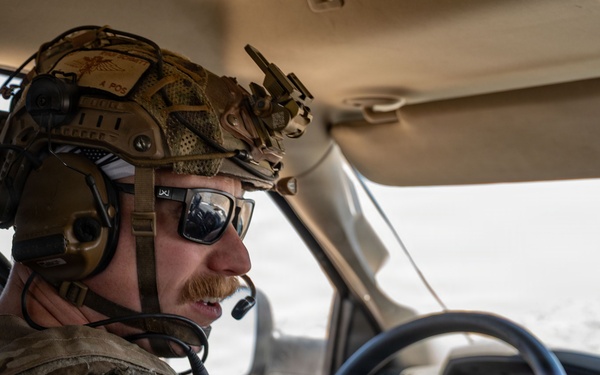 A military or tactical operator is driving a Ram pickup truck through what appears to be an arid, desert-like environment. The individual is wearing full tactical gear including a combat helmet with mounted accessories, communications headset with boom microphone, sunglasses, and a plate carrier or tactical vest. The vehicle interior and steering wheel are clearly visible, with the Ram logo prominently displayed on the steering wheel.