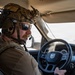 A military or tactical operator is driving a Ram pickup truck through what appears to be an arid, desert-like environment. The individual is wearing full tactical gear including a combat helmet with mounted accessories, communications headset with boom microphone, sunglasses, and a plate carrier or tactical vest. The vehicle interior and steering wheel are clearly visible, with the Ram logo prominently displayed on the steering wheel.