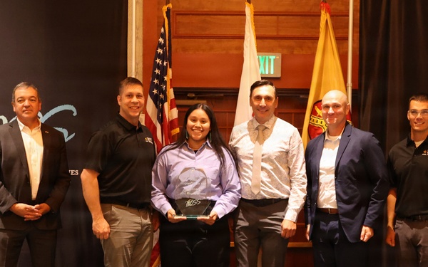 A group of six individuals pose on a stage at what appears to be a U.S. Army BOSS (Better Opportunities for Single Soldiers) award ceremony. A young woman in the center is holding a plaque or award, surrounded by five men in business or semi-formal attire. The backdrop features U.S. Army and BOSS program branding, along with American and military flags.