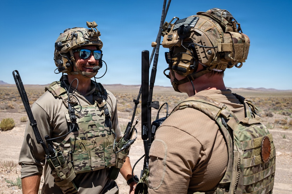 Two military personnel in full tactical gear are conversing in an arid desert environment under clear blue skies. Both individuals are equipped with helmets, communication headsets, plate carriers, and visible antennas suggesting active radio communication equipment. The setting appears to be a training or operational area in a desert region.