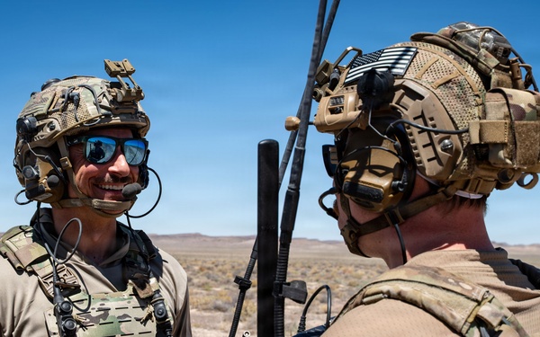 Two military personnel in full tactical gear are conversing in an arid desert environment under clear blue skies. Both individuals are equipped with helmets, communication headsets, plate carriers, and visible antennas suggesting active radio communication equipment. The setting appears to be a training or operational area in a desert region.