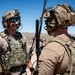 Two military personnel in full tactical gear are conversing in an arid desert environment under clear blue skies. Both individuals are equipped with helmets, communication headsets, plate carriers, and visible antennas suggesting active radio communication equipment. The setting appears to be a training or operational area in a desert region.
