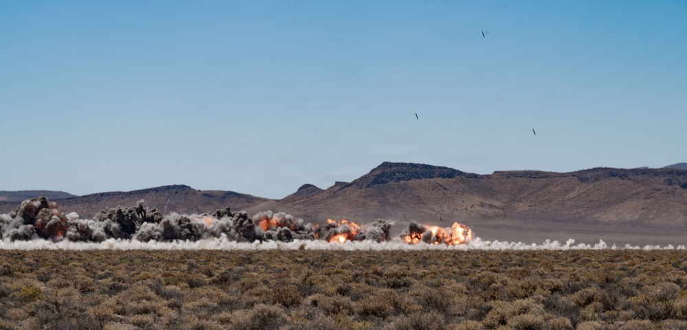 The image captures multiple simultaneous explosions occurring across a desert landscape, with large plumes of smoke, dust, and fire visible along a horizontal line. The setting appears to be an arid, scrubland terrain with mountain ridges visible in the background under a clear blue sky. This appears to be a military ordnance detonation or weapons testing exercise.