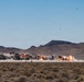The image captures multiple simultaneous explosions occurring across a desert landscape, with large plumes of smoke, dust, and fire visible along a horizontal line. The setting appears to be an arid, scrubland terrain with mountain ridges visible in the background under a clear blue sky. This appears to be a military ordnance detonation or weapons testing exercise.