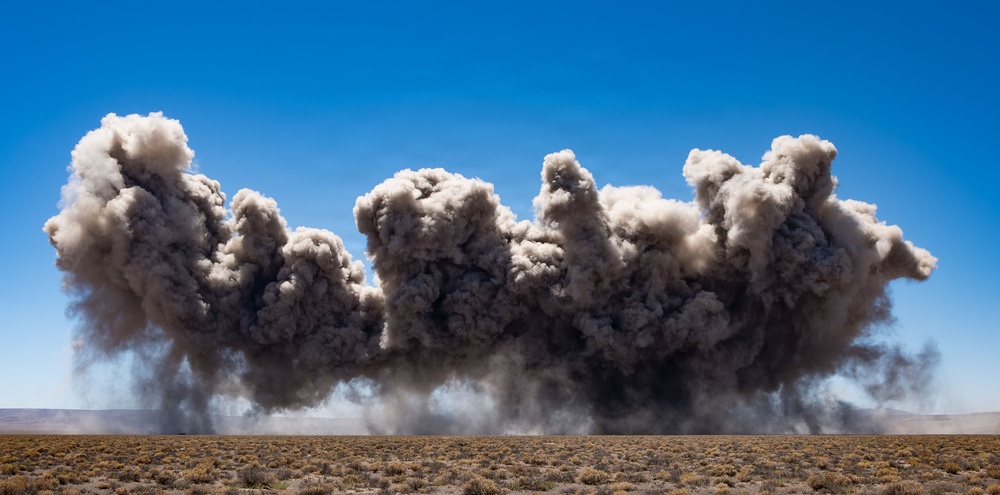A large dust and debris cloud rises horizontally across an arid desert landscape under a clear blue sky. The cloud appears to result from an explosion or controlled detonation at ground level, spreading laterally across the frame. The terrain is flat and sparse with low desert scrub vegetation.