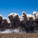 A large dust and debris cloud rises horizontally across an arid desert landscape under a clear blue sky. The cloud appears to result from an explosion or controlled detonation at ground level, spreading laterally across the frame. The terrain is flat and sparse with low desert scrub vegetation.