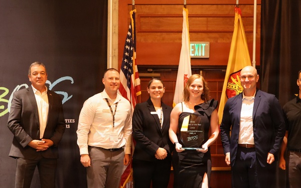 Six individuals are posed on a stage at a U.S. Army BOSS (Better Opportunities for Single Soldiers) event, with one person holding a crystal/glass award plaque. The backdrop features the U.S. Army star logo and BOSS branding, along with American and military flags. This appears to be an awards ceremony recognizing achievement within the BOSS program.