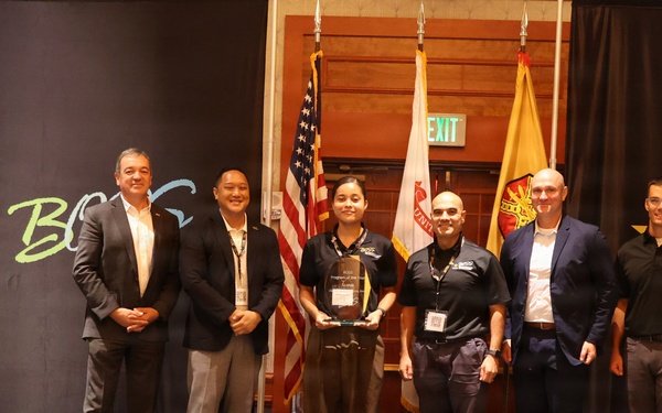 A group of six individuals pose on a stage at a U.S. Army BOSS (Better Opportunities for Single Soldiers) program event. One person in the center is holding a clear award or trophy, suggesting a recognition or award ceremony. The backdrop features U.S. Army and BOSS program branding, with American and military flags visible in the background.