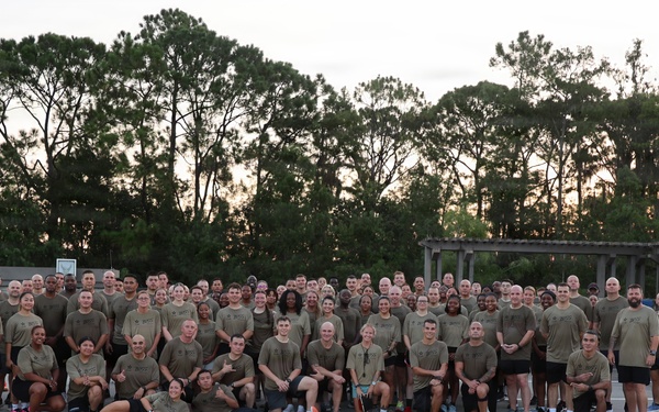 A large group of approximately 100-150 individuals dressed in matching olive/tan t-shirts and dark shorts pose for a group photo on what appears to be a rooftop parking structure. The setting suggests a military or law enforcement unit physical training (PT) event, likely taken at dawn or dusk given the lighting conditions. The group is arranged in multiple rows with some individuals kneeling or sitting in the front.