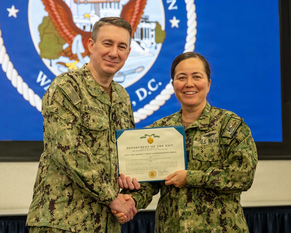 Two U.S. Navy personnel in Navy Working Uniform (NWU Type III) are posing together while holding a Department of the Navy certificate for the Navy and Marine Corps Commendation Medal. The male officer on the left, identified by his name tape as 'Lawrence,' is presenting the award to the female sailor on the right wearing a 'U.S. Navy' tape. The ceremony appears to be taking place in front of a projected Department of the Navy seal backdrop.