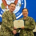Two U.S. Navy personnel in Navy Working Uniform (NWU Type III) are posing together while holding a Department of the Navy certificate for the Navy and Marine Corps Commendation Medal. The male officer on the left, identified by his name tape as 'Lawrence,' is presenting the award to the female sailor on the right wearing a 'U.S. Navy' tape. The ceremony appears to be taking place in front of a projected Department of the Navy seal backdrop.