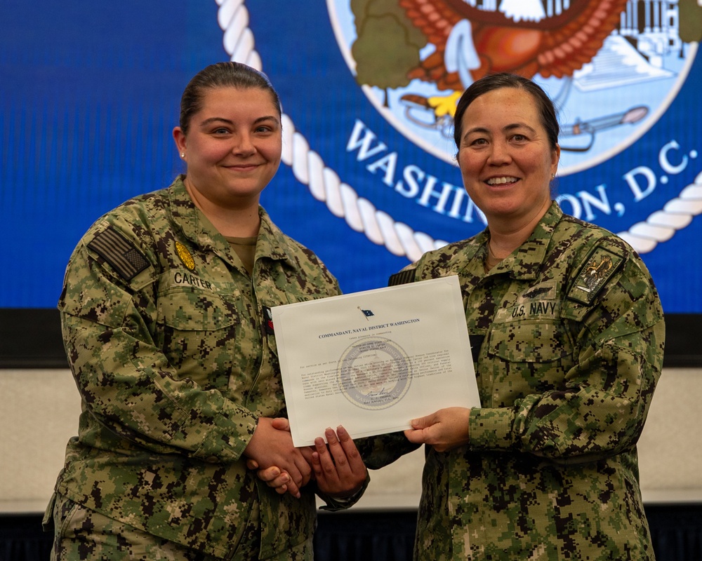 Two U.S. Navy personnel in NWU Type III (woodland digital camouflage) uniforms pose together while jointly holding a certificate from the Commandant, Naval District Washington. The individual on the left, identified by name tape as 'CARTER,' appears to be receiving the award. The ceremony is taking place in front of a large Naval District Washington, D.C. official seal displayed on a screen.
