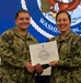 Two U.S. Navy personnel in NWU Type III (woodland digital camouflage) uniforms pose together while jointly holding a certificate from the Commandant, Naval District Washington. The individual on the left, identified by name tape as 'CARTER,' appears to be receiving the award. The ceremony is taking place in front of a large Naval District Washington, D.C. official seal displayed on a screen.