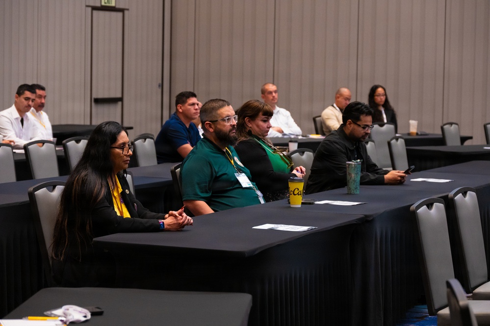 A group of approximately 10 attendees are seated at black-draped tables in a conference or meeting room, appearing to listen to a presentation or speaker outside the frame. Participants wear name badge lanyards and casual to business-casual attire, with some in matching green shirts suggesting organizational affiliation. The room appears to be a hotel or convention center ballroom with many empty chairs.