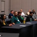 A group of approximately 10 attendees are seated at black-draped tables in a conference or meeting room, appearing to listen to a presentation or speaker outside the frame. Participants wear name badge lanyards and casual to business-casual attire, with some in matching green shirts suggesting organizational affiliation. The room appears to be a hotel or convention center ballroom with many empty chairs.