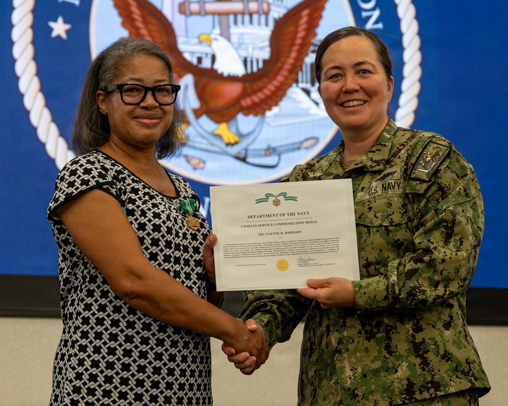 A civilian woman and a U.S. Navy officer are jointly holding a Department of the Navy Civilian Service Commendation Medal certificate while shaking hands. The civilian recipient, identified on the certificate as Ms. Yvette D. Johnson, is wearing the medal pinned to her dress. The ceremony appears to be taking place in front of a large U.S. Navy seal backdrop.