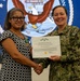 A civilian woman and a U.S. Navy officer are jointly holding a Department of the Navy Civilian Service Commendation Medal certificate while shaking hands. The civilian recipient, identified on the certificate as Ms. Yvette D. Johnson, is wearing the medal pinned to her dress. The ceremony appears to be taking place in front of a large U.S. Navy seal backdrop.