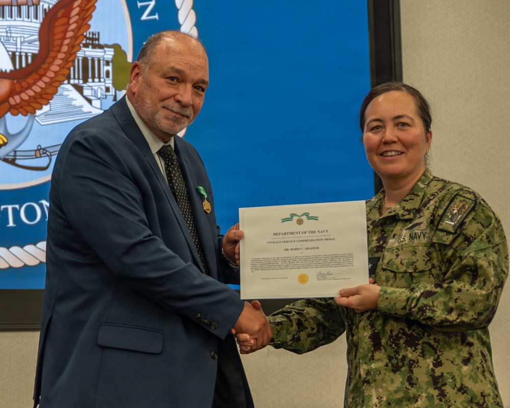 A U.S. Navy officer in digital camouflage uniform is presenting a Department of the Navy Civilian Service Commendation Medal certificate to a civilian man in a blue suit. The civilian recipient is already wearing the medal on his lapel. The ceremony appears to be taking place in an official Navy facility with a Navy seal displayed on a screen in the background.