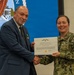 A U.S. Navy officer in digital camouflage uniform is presenting a Department of the Navy Civilian Service Commendation Medal certificate to a civilian man in a blue suit. The civilian recipient is already wearing the medal on his lapel. The ceremony appears to be taking place in an official Navy facility with a Navy seal displayed on a screen in the background.
