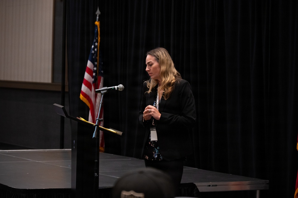 A woman in a black blazer stands at a podium with a microphone, appearing to speak or present at a formal event. She is wearing a lanyard with a badge and is looking downward, possibly at notes. An American flag is visible in the background against black curtain draping.