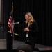 A woman in a black blazer stands at a podium with a microphone, appearing to speak or present at a formal event. She is wearing a lanyard with a badge and is looking downward, possibly at notes. An American flag is visible in the background against black curtain draping.