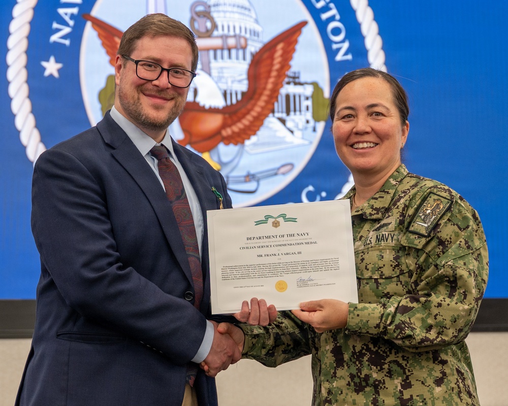 A civilian male and a U.S. Navy uniformed officer are jointly holding a Department of the Navy Civilian Service Commendation Medal certificate awarded to Mr. Frank J. Vargas, III. Both individuals are smiling and shaking hands in front of a large Navy seal backdrop. The setting appears to be a formal military awards ceremony.