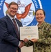 A civilian male and a U.S. Navy uniformed officer are jointly holding a Department of the Navy Civilian Service Commendation Medal certificate awarded to Mr. Frank J. Vargas, III. Both individuals are smiling and shaking hands in front of a large Navy seal backdrop. The setting appears to be a formal military awards ceremony.