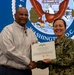 A U.S. Navy officer in digital camouflage uniform presents a Department of the Navy Civilian Service Commendation Medal certificate to a civilian man named Mr. Edward C. Zeigler. Both individuals are smiling and jointly holding the certificate for the photo. The ceremony appears to take place in front of a Naval District Washington, D.C. seal backdrop.