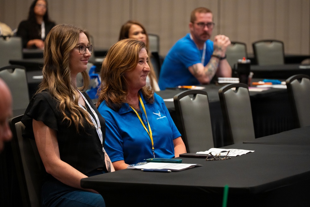 The image shows attendees seated at tables during what appears to be a professional conference or training session. Two women in the foreground are the primary focus, one wearing a blue polo shirt with a yellow lanyard and the other in a black top with a white lanyard. Additional attendees are visible in the background, including a man in a blue shirt with visible tattoos.