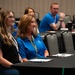 The image shows attendees seated at tables during what appears to be a professional conference or training session. Two women in the foreground are the primary focus, one wearing a blue polo shirt with a yellow lanyard and the other in a black top with a white lanyard. Additional attendees are visible in the background, including a man in a blue shirt with visible tattoos.