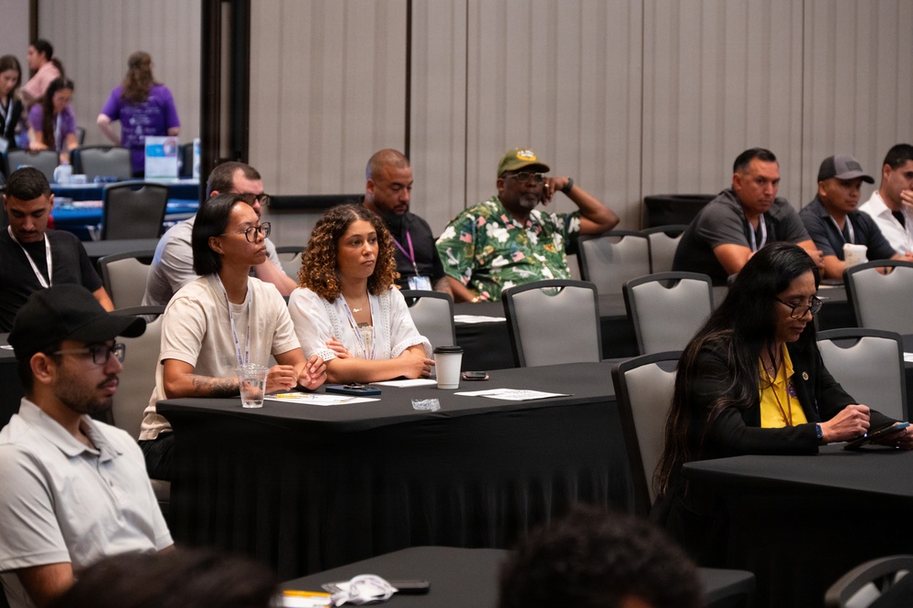 A conference or seminar session is taking place in a large meeting room with attendees seated at round tables covered with black tablecloths. Participants appear to be listening to a presentation, with some taking notes or using devices. The room is partially filled with a diverse group of attendees, some wearing lanyards and name badges.