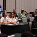 A conference or seminar session is taking place in a large meeting room with attendees seated at round tables covered with black tablecloths. Participants appear to be listening to a presentation, with some taking notes or using devices. The room is partially filled with a diverse group of attendees, some wearing lanyards and name badges.