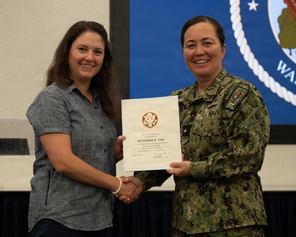 A U.S. Navy officer in NWU Type III camouflage uniform is presenting a certificate to a civilian woman during a formal recognition ceremony. The certificate is awarded to Katherine N. Tice in recognition of fifteen years of service to the Government of the United States of America. Both individuals are smiling and shaking hands while jointly holding the certificate.