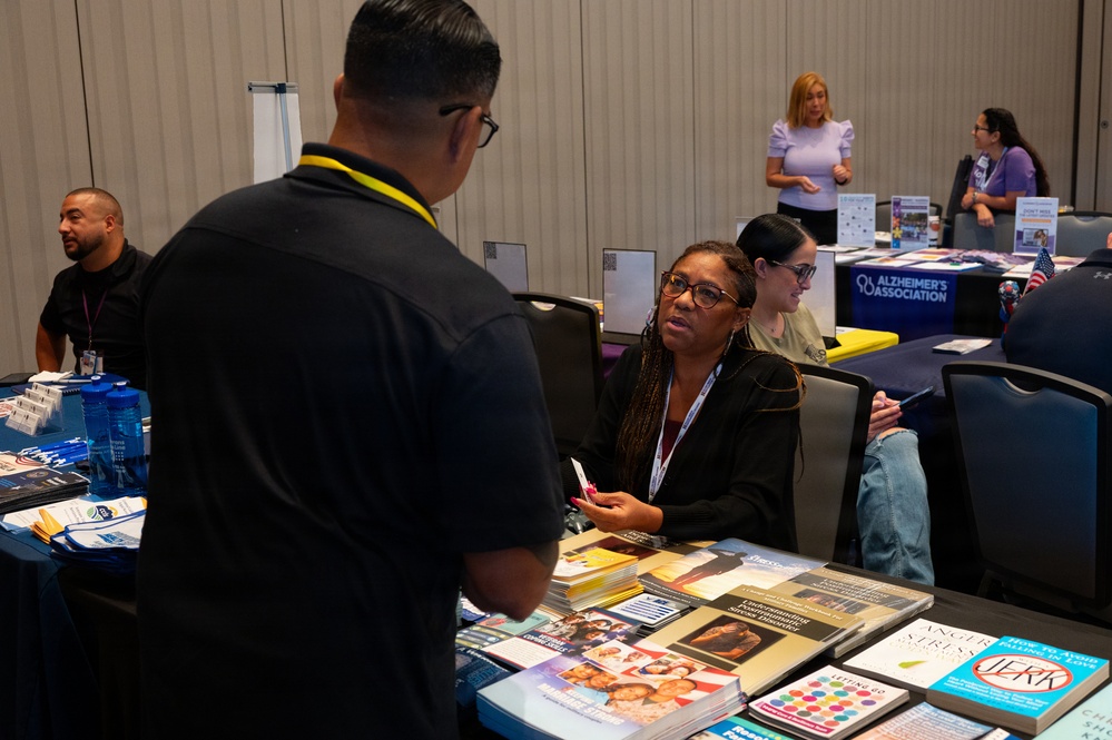 The image shows a health or community resource fair taking place in a conference or convention hall. Multiple vendor tables are set up with informational materials, books, and brochures, including a visible Alzheimer's Association booth in the background. Attendees and representatives are engaged in conversations at various tables.