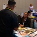 The image shows a health or community resource fair taking place in a conference or convention hall. Multiple vendor tables are set up with informational materials, books, and brochures, including a visible Alzheimer's Association booth in the background. Attendees and representatives are engaged in conversations at various tables.