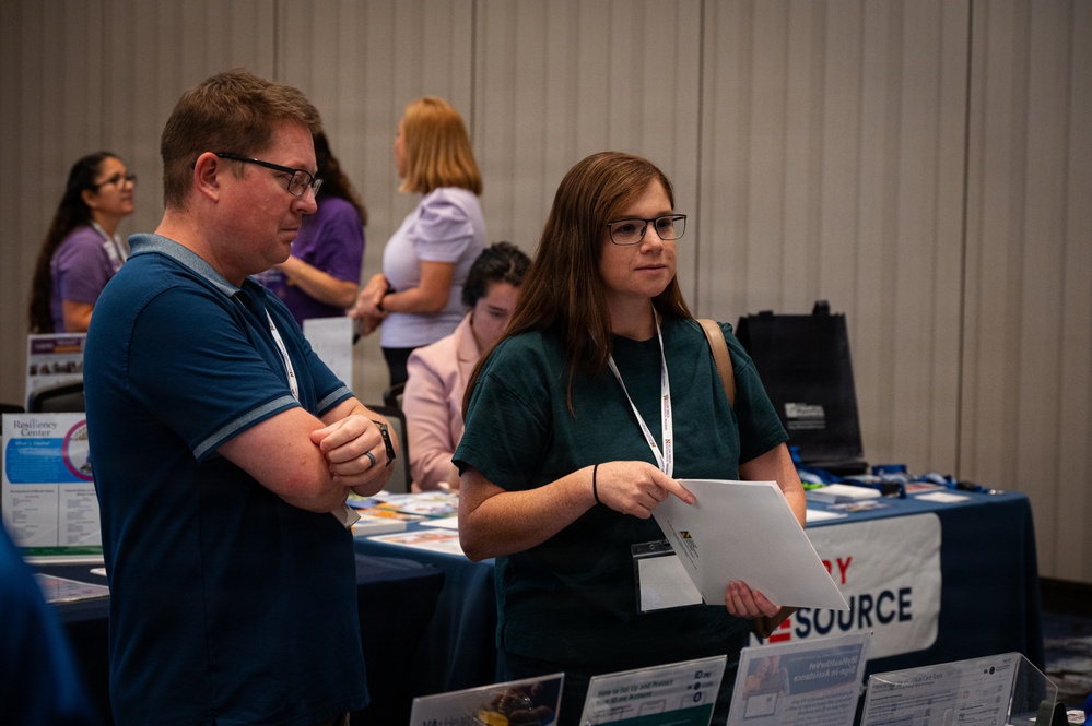 The image shows attendees at what appears to be a conference or resource fair, browsing informational tables with brochures and displays. Two people in the foreground — a man in a blue polo shirt and a woman in a teal shirt holding papers — appear to be reviewing available resources. Several other attendees and booth representatives are visible in the background.