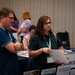 The image shows attendees at what appears to be a conference or resource fair, browsing informational tables with brochures and displays. Two people in the foreground — a man in a blue polo shirt and a woman in a teal shirt holding papers — appear to be reviewing available resources. Several other attendees and booth representatives are visible in the background.