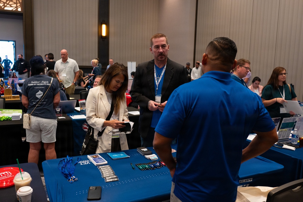 The image depicts a busy indoor expo or job fair event held in a large conference hall. Multiple vendor tables are set up with promotional materials, lanyards, and brochures, and attendees are actively engaging with representatives. One table in the foreground prominently displays blue tablecloths with items labeled 'VET' suggesting a veterans-focused event.