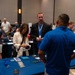 The image depicts a busy indoor expo or job fair event held in a large conference hall. Multiple vendor tables are set up with promotional materials, lanyards, and brochures, and attendees are actively engaging with representatives. One table in the foreground prominently displays blue tablecloths with items labeled 'VET' suggesting a veterans-focused event.