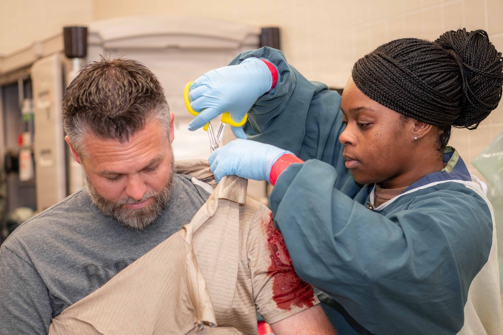 A medical professional wearing blue latex gloves is using trauma shears (scissors) to cut away clothing from a patient's upper arm/shoulder area, which shows visible simulated blood. The patient, a middle-aged man with gray hair and a beard, appears to be in a medical or training facility. The scene is consistent with emergency medical care or a trauma training exercise.