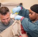 A medical professional wearing blue latex gloves is using trauma shears (scissors) to cut away clothing from a patient's upper arm/shoulder area, which shows visible simulated blood. The patient, a middle-aged man with gray hair and a beard, appears to be in a medical or training facility. The scene is consistent with emergency medical care or a trauma training exercise.