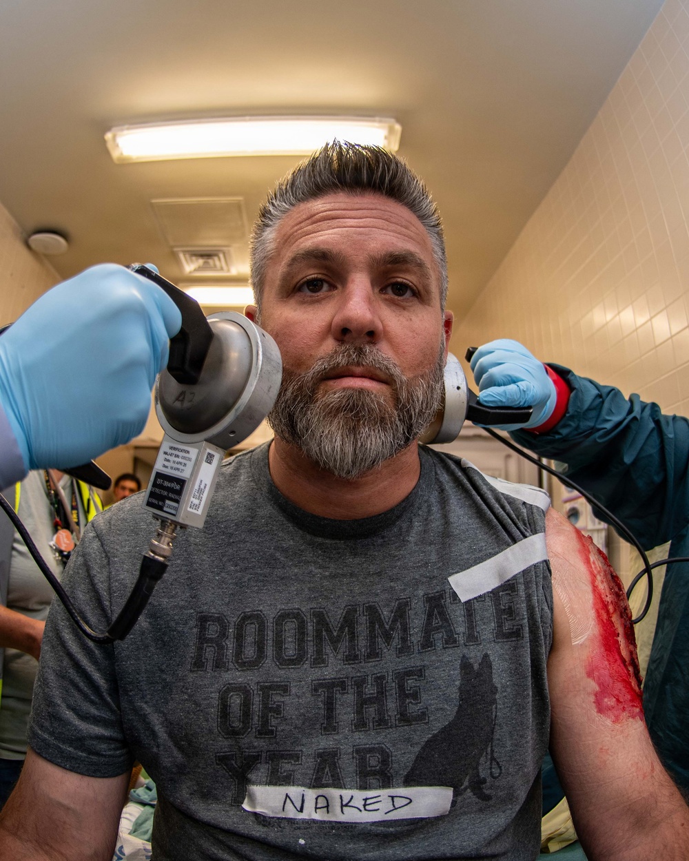 A middle-aged man with gray hair and a beard is seated while two individuals wearing blue medical/examination gloves hold devices to each side of his head. He is wearing a gray t-shirt reading 'Roommate of the Year Naked' with a dog graphic. A visibly bloody wound or simulated injury is visible on his right arm/shoulder area.