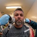 A middle-aged man with gray hair and a beard is seated while two individuals wearing blue medical/examination gloves hold devices to each side of his head. He is wearing a gray t-shirt reading 'Roommate of the Year Naked' with a dog graphic. A visibly bloody wound or simulated injury is visible on his right arm/shoulder area.